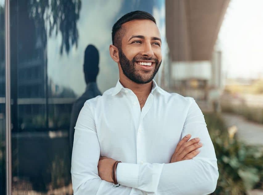 Smile Makeover page content image 2 A man with a short beard standing outside wearing a white button-up with arms folded smiling